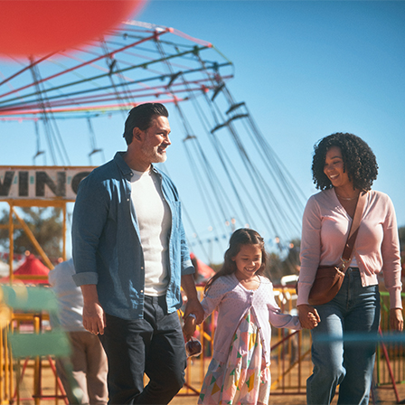 father and mother holding hands of daughter with swing ride in the background