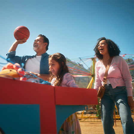 father playing basketball game at carnival while mother and daughter smile and watch