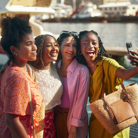 four women taking a selfie with body of water and boats behind them