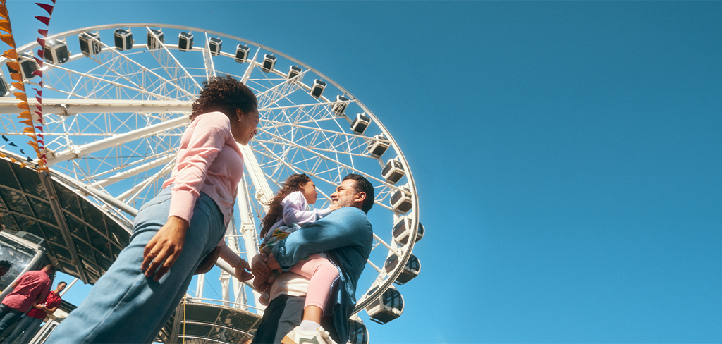 Madre mirando al padre con su hija en brazos en una feria, con la rueda de la fortuna en el fondo.